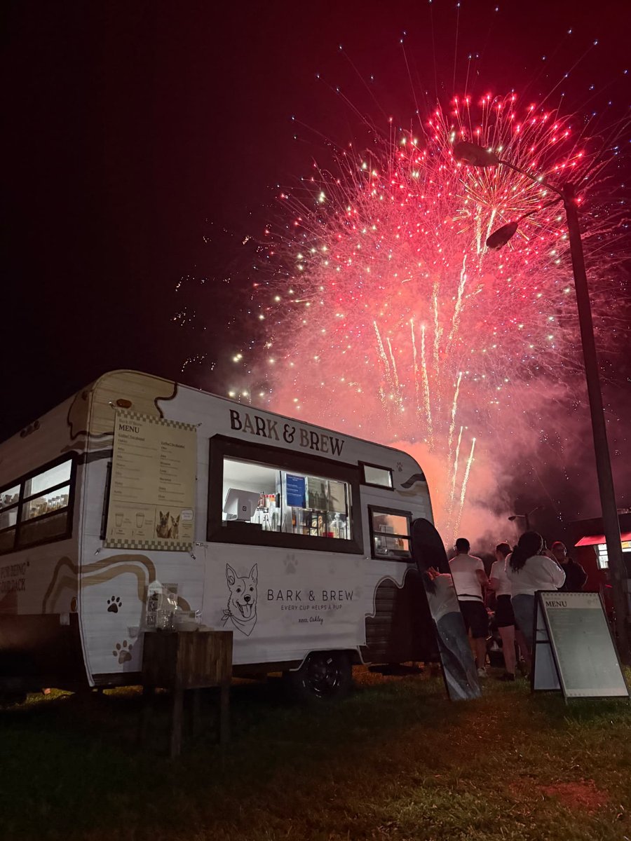 The lit-up Bark & Brew trailer at night with guests outside and fireworks overhead