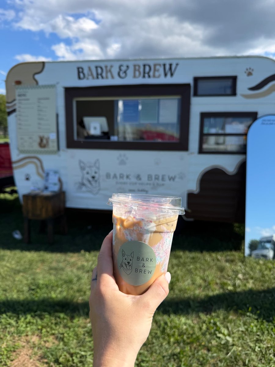 An iced coffee held up in front of the Bark & Brew trailer parked on the grass