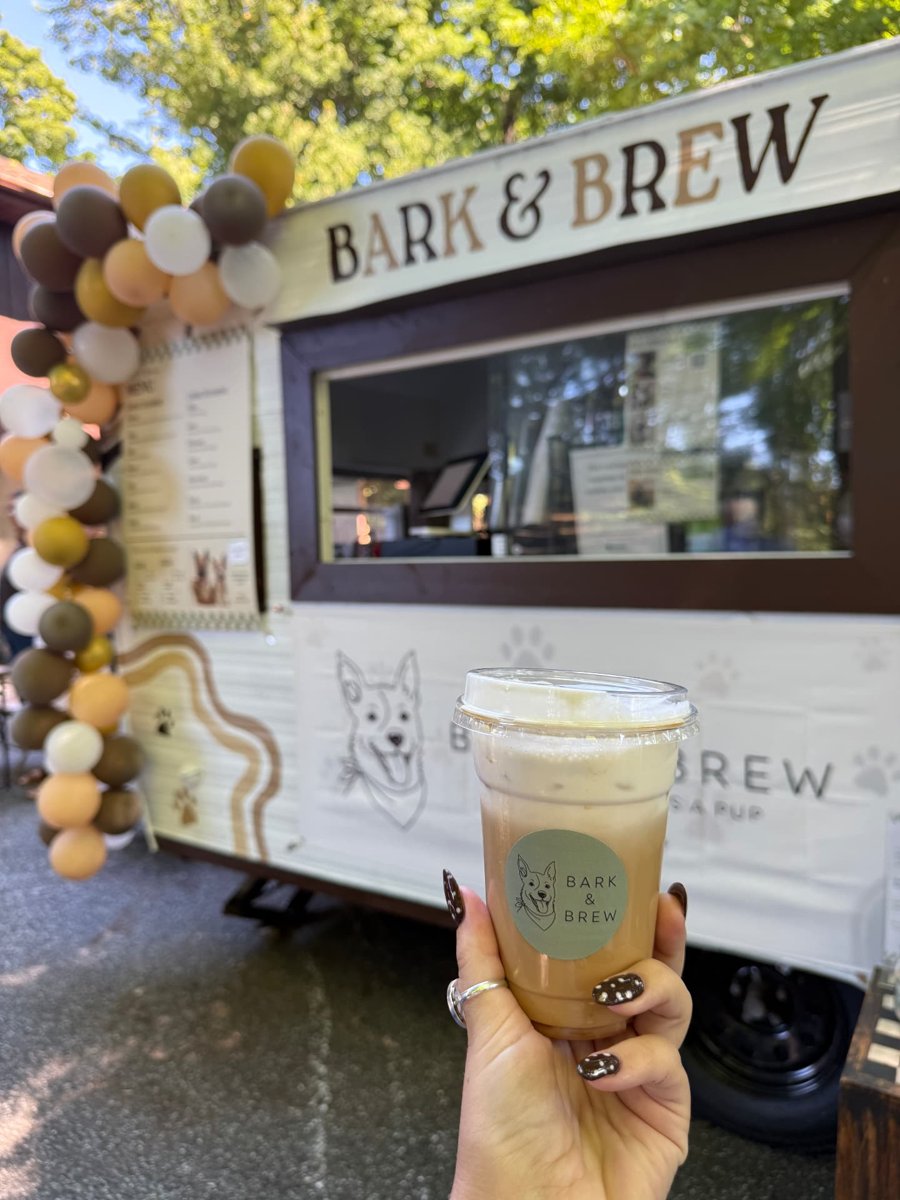 A hand holding a Bark & Brew iced coffee in front of the decorated trailer
